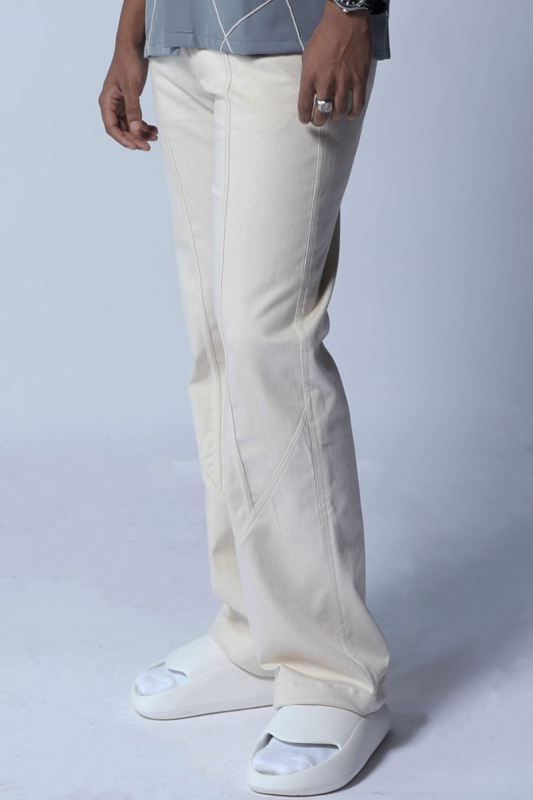 Close-up of a person wearing cream-colored straight-leg pants with front crease detail, paired with white slip-on sandals and a casual grey printed top, standing against a plain light background