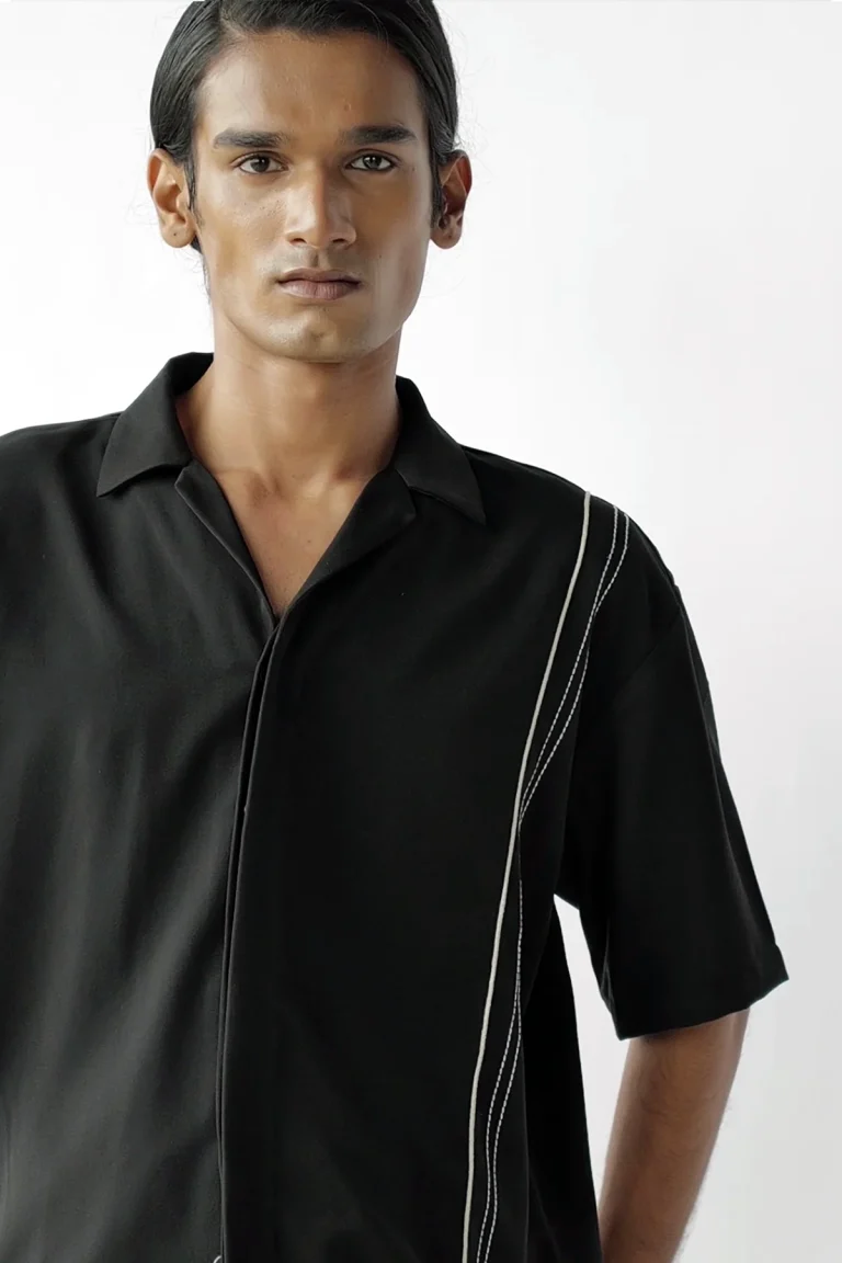 A person with straight dark hair wears a black shirt for men featuring white diagonal stitch lines, standing against a plain light background.