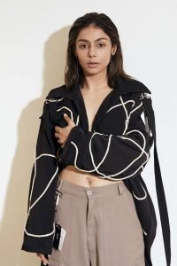 Young woman with long hair stands against a white background. She wears a black corded jacket with white abstract lines and beige pants.