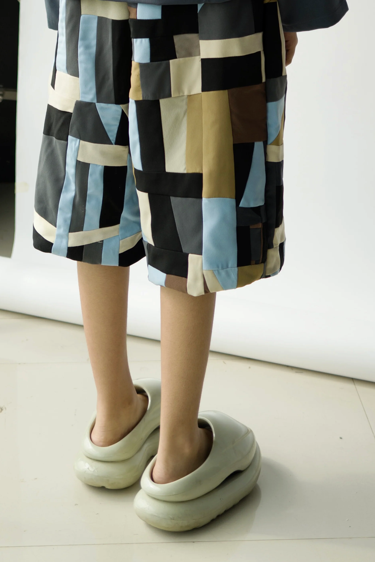 A person wearing oversized patchwork shorts with abstract patterns stands indoors, facing away.