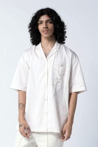 Young person with curly hair wearing a loose, white old money shirt and white pants stands confidently against a plain backdrop.